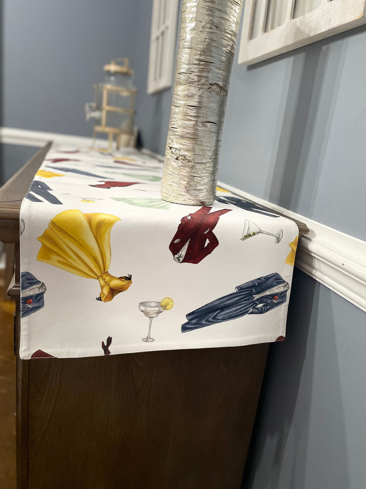 The image shows a wooden table or sideboard with a decorative runner draped over it. The runner has a vibrant pattern featuring images of dresses and suits from the 1950’s, along with martini glasses. Sophisticated and stylish decor theme.