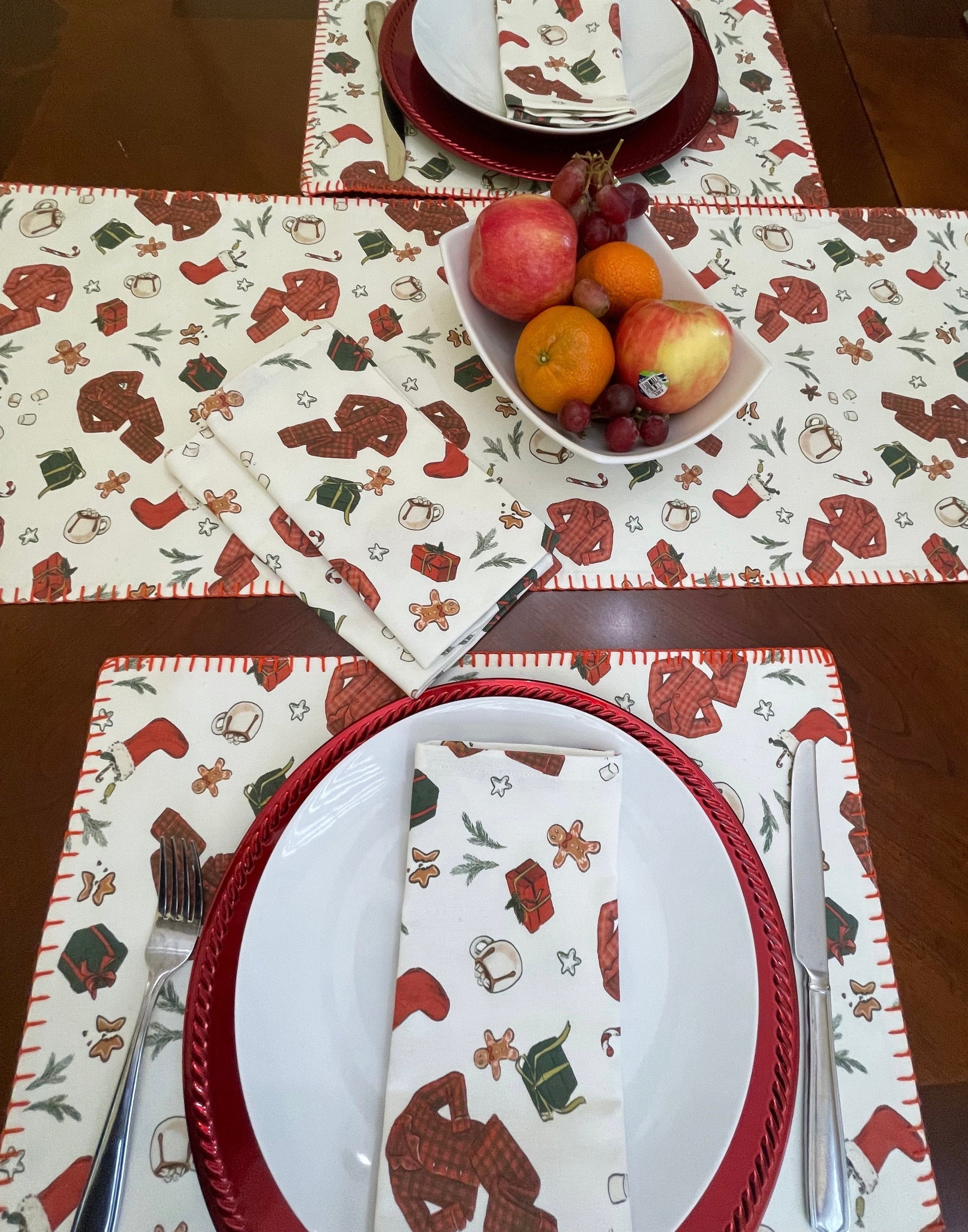 Set table with Christmas-themed placemats, napkins, and a bowl of fruit on a wooden table.