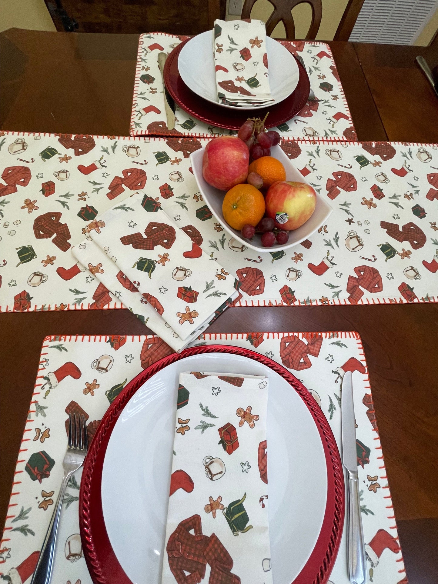Anghild's Table Placemat Christmas Morning Placemats set of 2 Christmas Morning Placemats set of 2 Anghild's Table Christmas Morning Cotton placemat with chritmas morning motif of buffalo check PJs, candy canes, Christmas  stockings, wrapped gifts and Christmas cookies on an off white background and red whip sticking trim. Matching cotton napkins on plate.